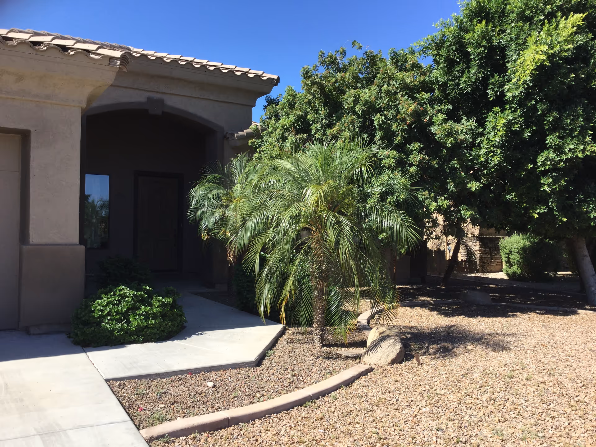 Exterior view of a single-story building entrance with a covered porch, surrounded by desert landscaping including palm trees, bushes, and gravel. The sky is clear and blue.