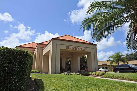 Sunlit building entrance labeled Aristocrat with red-tiled roof, palm trees, and parked cars nearby.