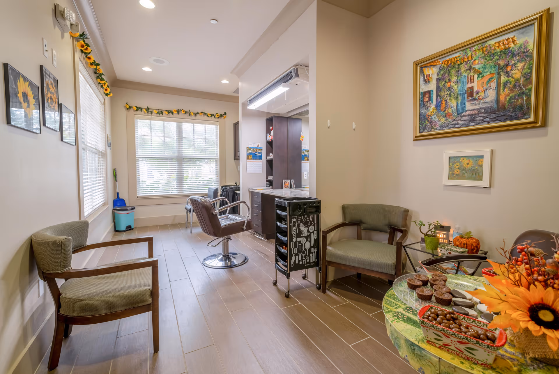 Interior salon/waiting area with a styling chair, waiting chairs, a rolling cart, and a snack table decorated with flowers and artwork.