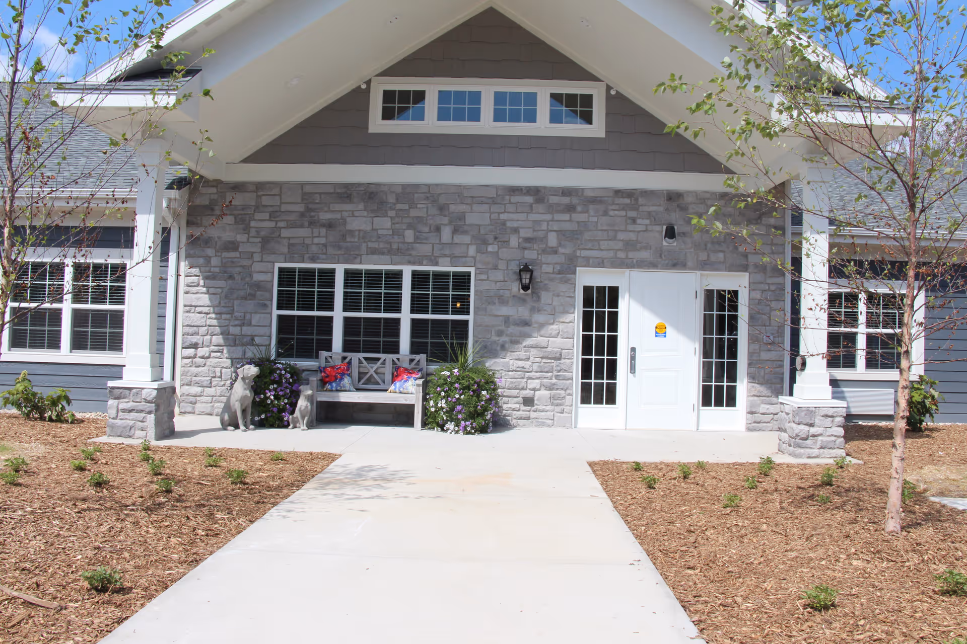 Front entrance of a senior living facility with a stone facade, white double doors with glass panels, a bench with decorative pillows, two dog statues, and small landscaped plants and trees on either side of a concrete walkway.