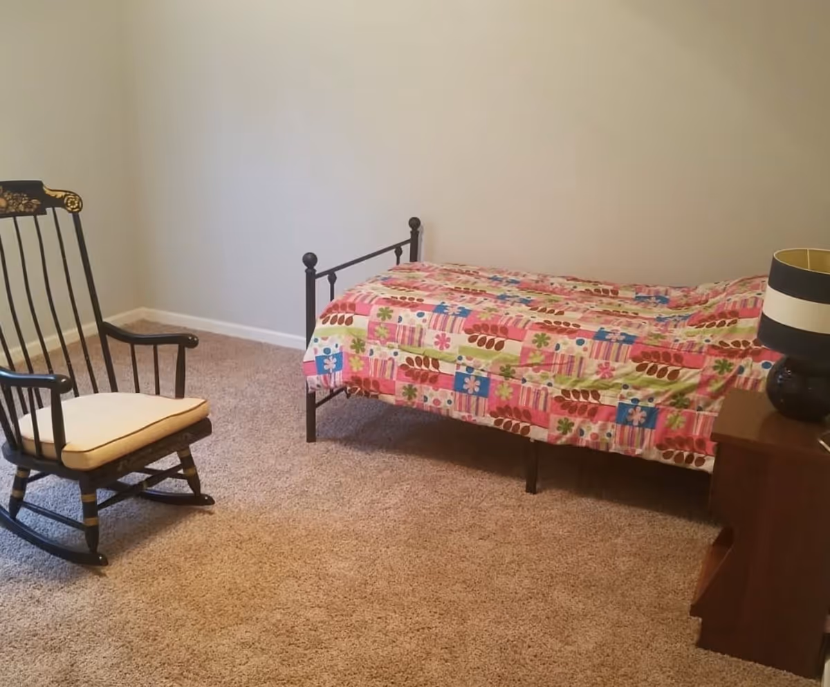 A simple bedroom with beige carpet, a single bed with a colorful patchwork quilt, a wooden nightstand with a black and white striped lamp, and a black wooden rocking chair with a white cushion.