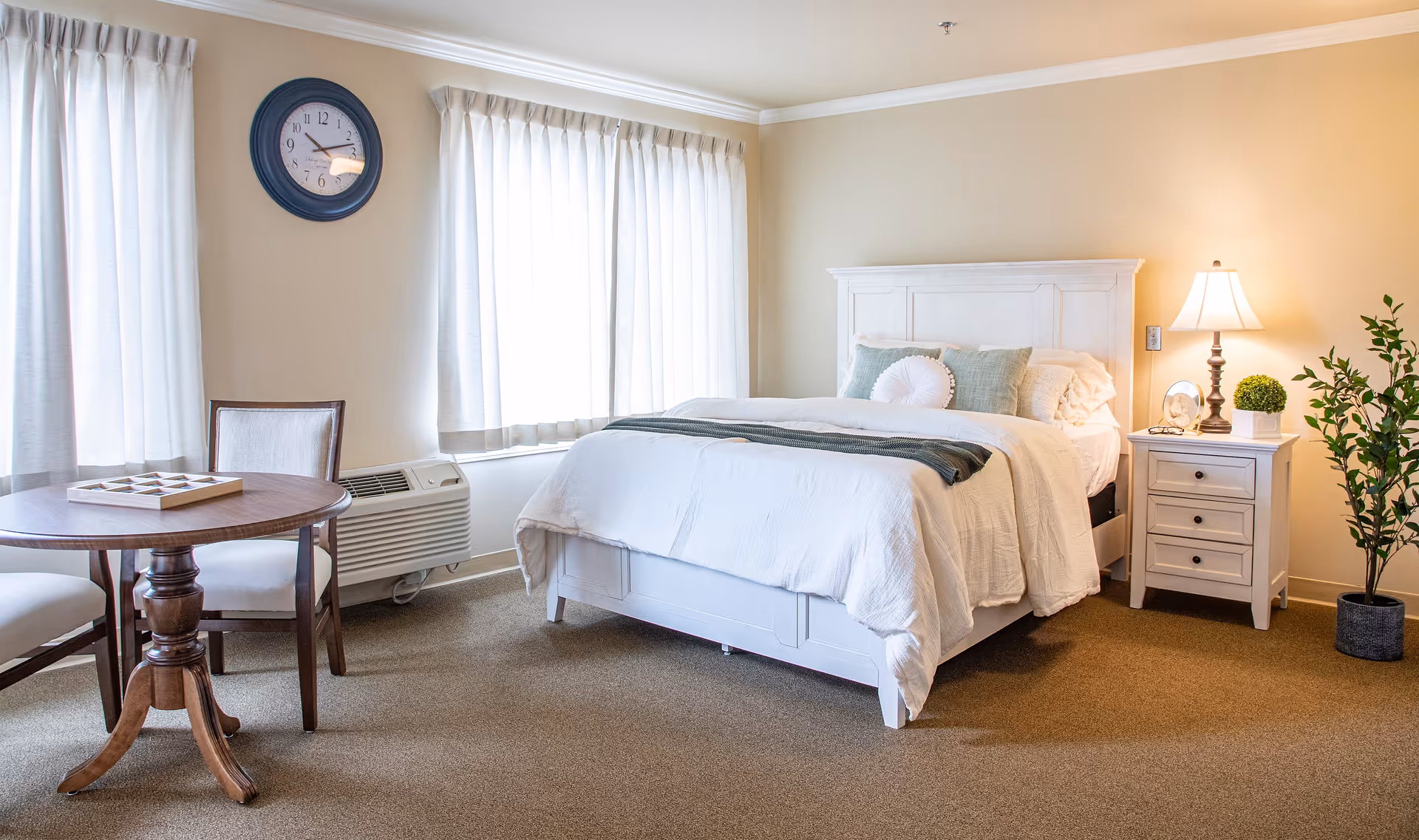 A cozy bedroom with a white wooden bed frame, white bedding, and decorative pillows. Next to the bed is a white nightstand with a lamp, a small plant, and a framed photo. There is a round wooden table with two chairs near two windows covered with white curtains. A wall clock is mounted above the windows, and a potted plant is placed on the floor near the nightstand.