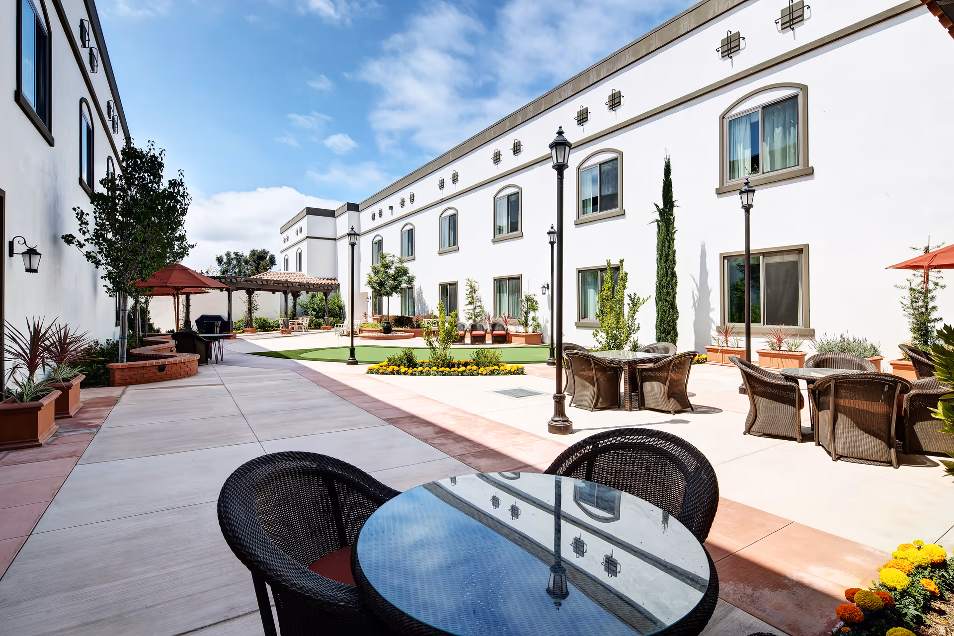 Outdoor courtyard area at Clearwater at South Bay featuring round glass-top tables with wicker chairs, potted plants, lamp posts, and a covered seating area with a red umbrella. The courtyard is surrounded by a white two-story building with multiple windows and a clear blue sky above.