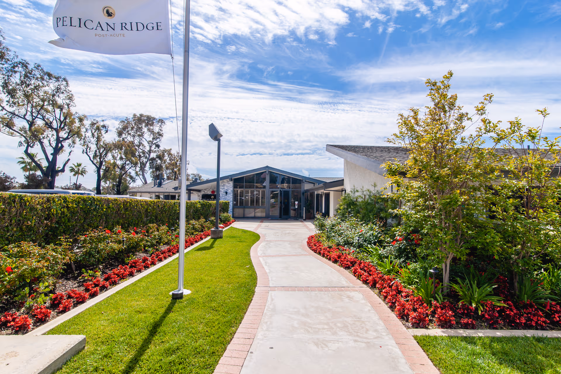 A paved walkway lined with red flowers and green shrubs leads to the entrance of a building under a partly cloudy sky. A flagpole with a white flag that reads 'Pelican Ridge Post-Acute' is on the left side of the walkway.