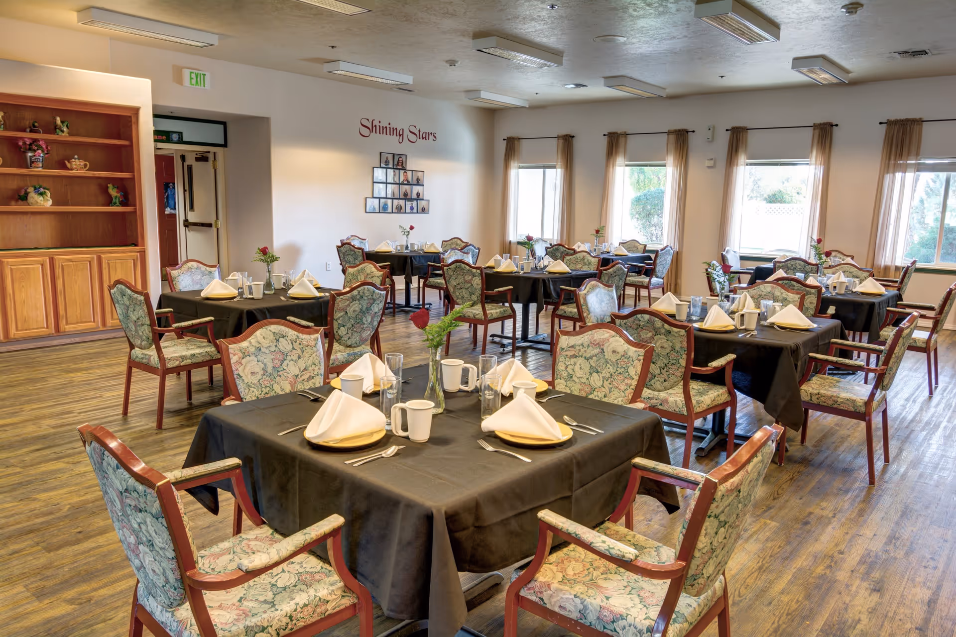 A dining room with multiple tables covered in black tablecloths, each set with white napkins, plates, cups, and silverware. The chairs have floral upholstery and wooden frames. The room has large windows with sheer curtains allowing natural light to enter. A wall features a display titled 'Shining Stars' with photos arranged in a pyramid shape. Wooden shelves and cabinets are visible on one side of the room.