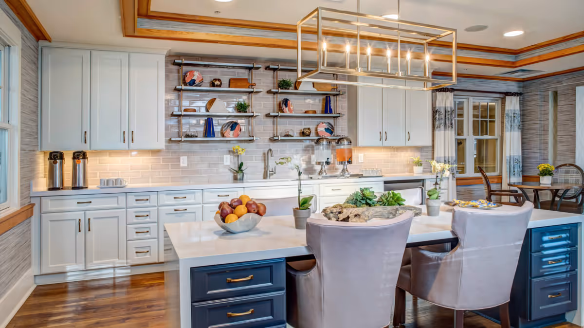 Bright modern kitchen with white cabinets, a large island with upholstered chairs, open shelving and a rectangular pendant light.