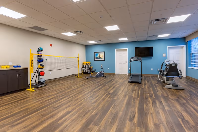 Spacious, well-lit fitness room with wood-look flooring containing a treadmill, recumbent bike, stationary bike, a small volleyball net, medicine balls, and a wall-mounted TV.