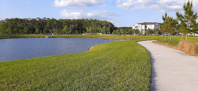 A paved walking path curves alongside a grassy area next to a pond with trees in the background and a multi-story building visible in the distance under a partly cloudy sky.