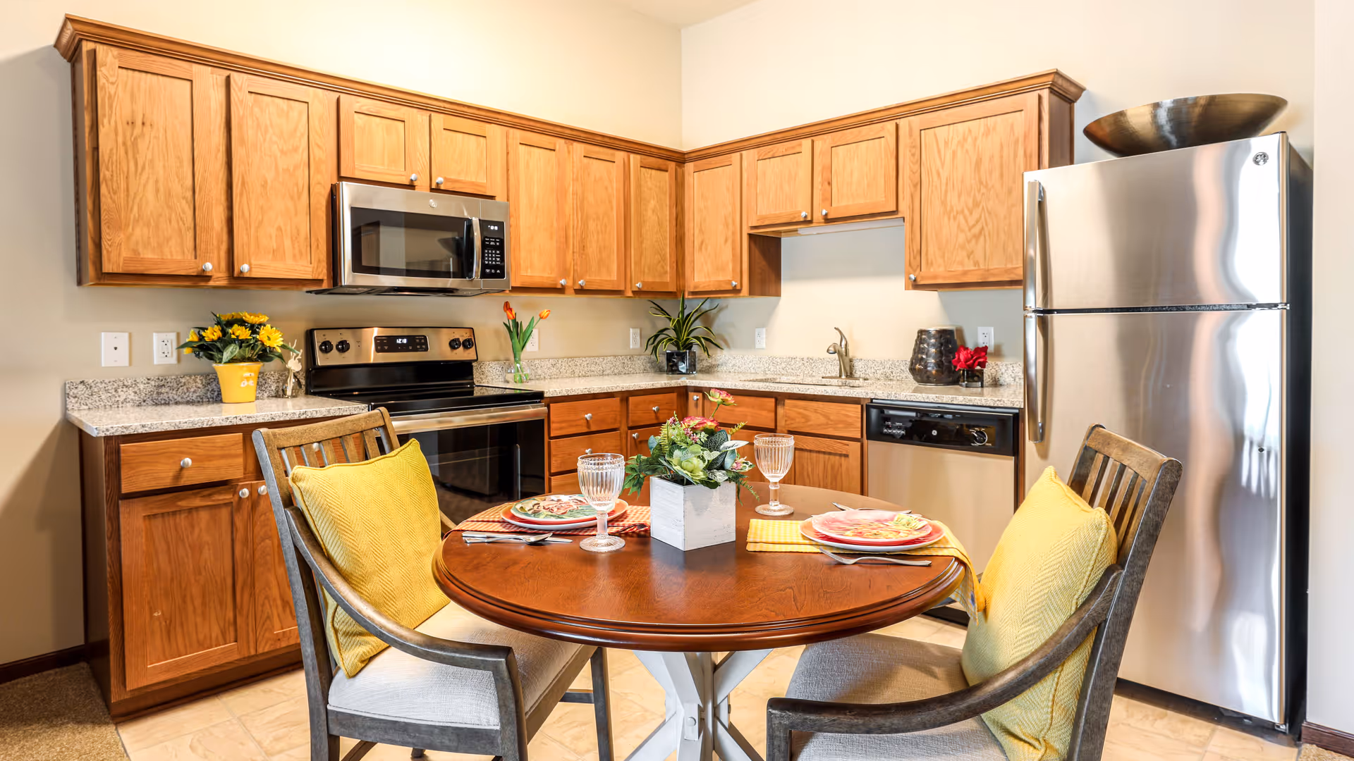 A bright kitchen with wooden cabinets, stainless steel appliances including a refrigerator, stove, microwave, and dishwasher. In the foreground, there is a round wooden dining table set for two with plates, glasses, and yellow cushions on the chairs. The countertop has decorative plants and flowers.