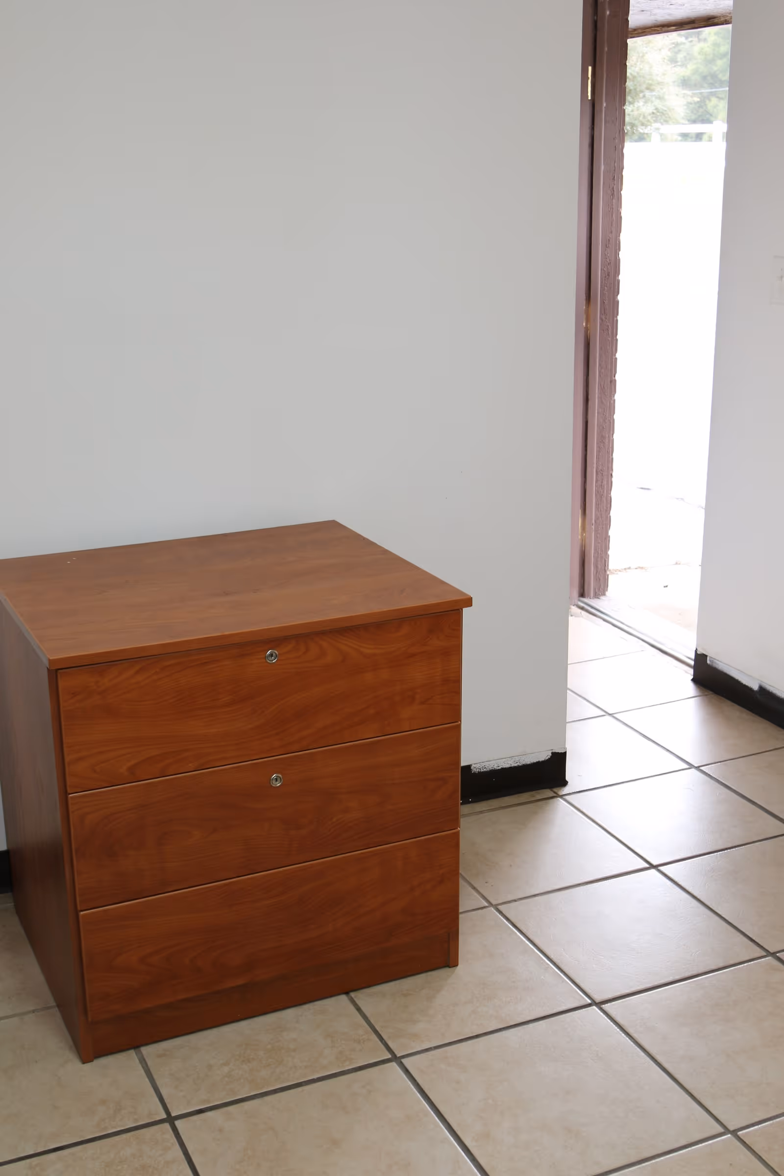A wooden three-drawer filing cabinet placed on a tiled floor next to a white wall and an open door leading outside.
