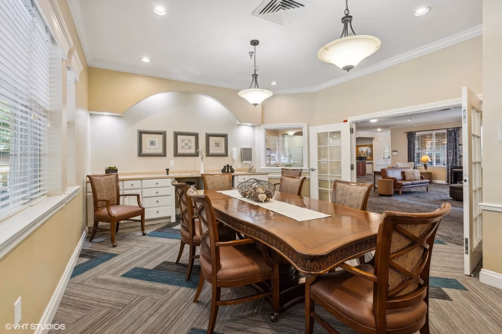 A well-lit dining room with a large wooden dining table surrounded by eight chairs. The room features beige walls, a patterned carpet with blue and gray tones, and three framed artworks on the far wall above a built-in cabinet with drawers. Two pendant lights hang from the ceiling. Through an open double door, a living room area with sofas and a window with curtains is visible.