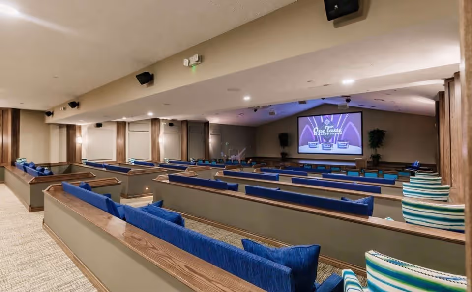 Interior view of a theater-style room with multiple rows of wooden benches featuring blue cushions and pillows. At the front, a large screen displays an advertisement for One Taste ice cream. The room has beige walls, carpeted floor, and ceiling-mounted speakers and lights.