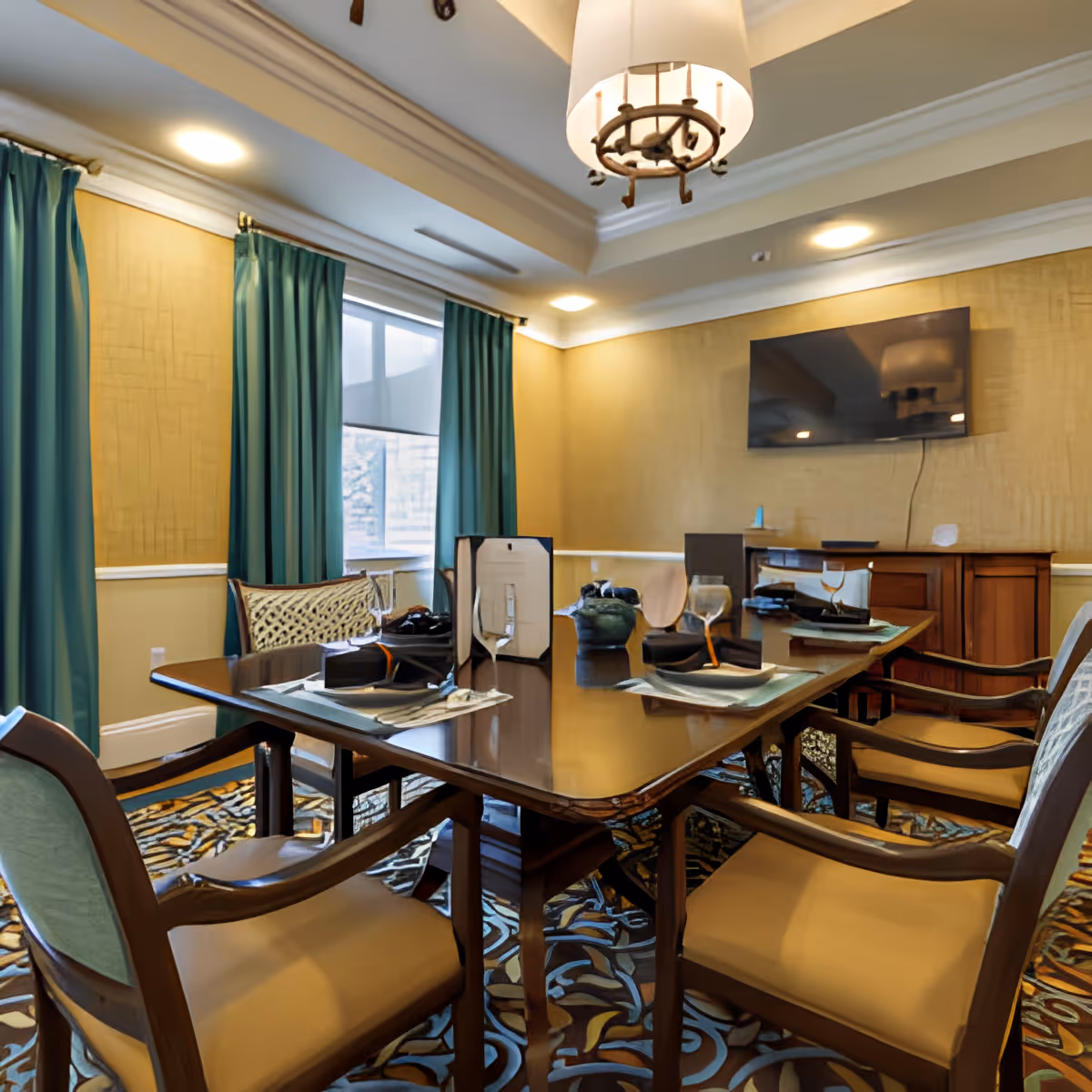 A formal dining room with a polished wooden table set for six, featuring wine glasses, napkins, and place settings. The room has teal curtains, a patterned carpet, a wall-mounted flat-screen TV, and warm yellow walls with crown molding. A chandelier hangs from the ceiling.