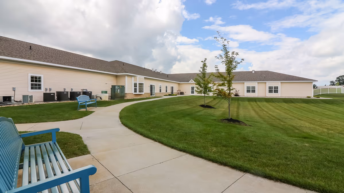 Paved pathway curves through a grassy courtyard with benches and young trees beside a single-story assisted living building under a cloudy sky.