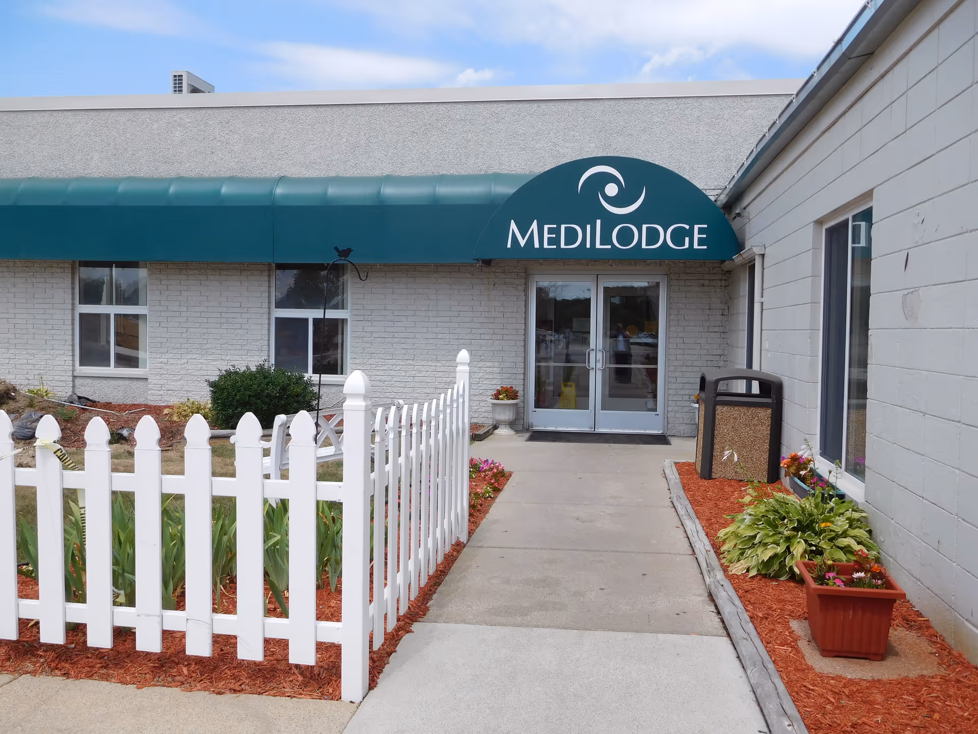 Front entrance of Medilodge with a green awning, double glass doors, a white picket fence, and landscaped walkway.