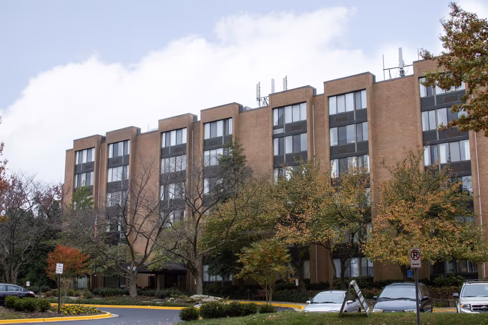 Front exterior of a multi-story brick senior living building with trees, landscaping, and parked cars at the entrance.