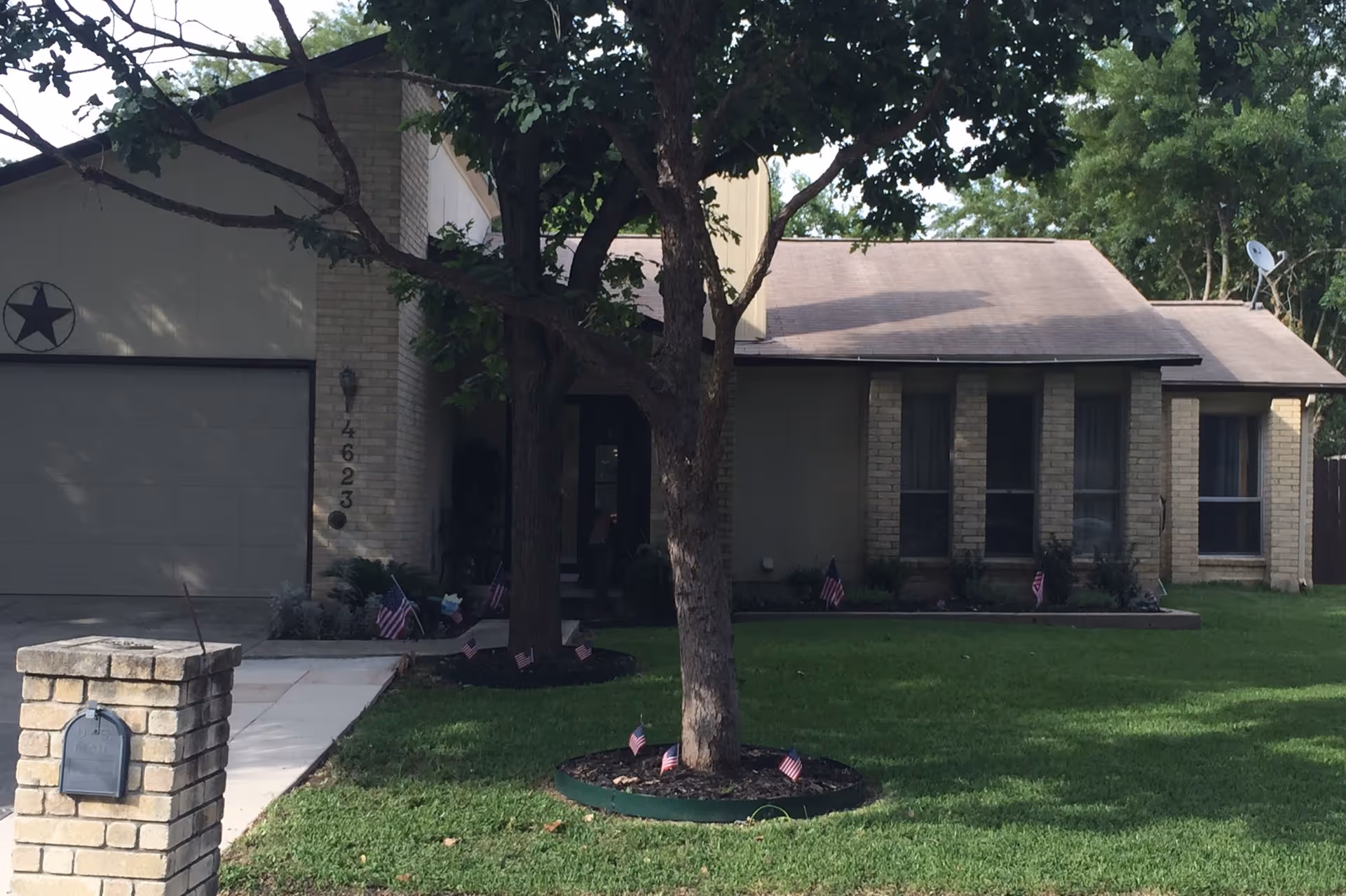 Front exterior of a single-story brick house with a tree, driveway, mailbox and small American flags on the lawn.