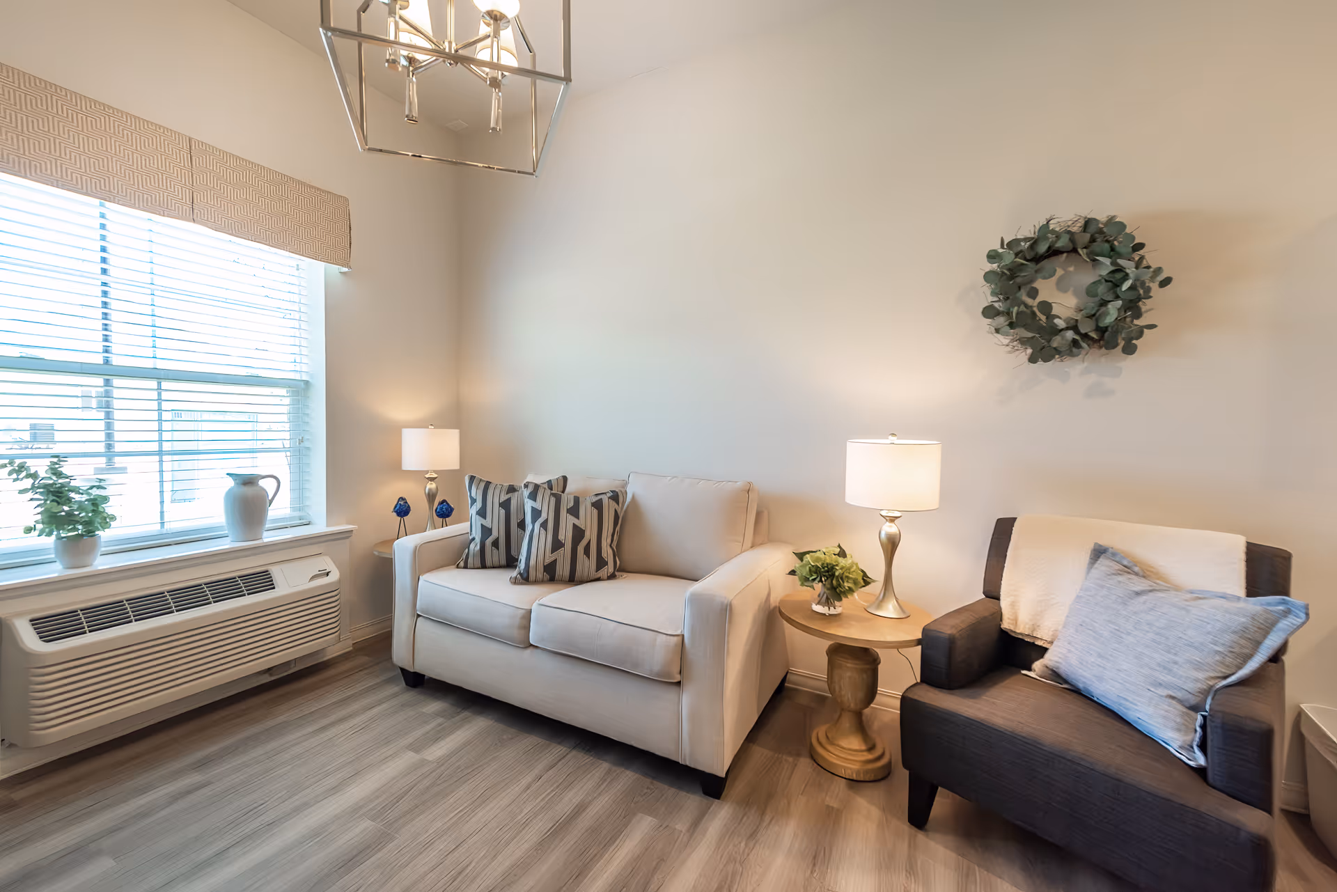 Bright living room with a beige loveseat, dark armchair, side table with lamp, window, and decorative wall wreath.
