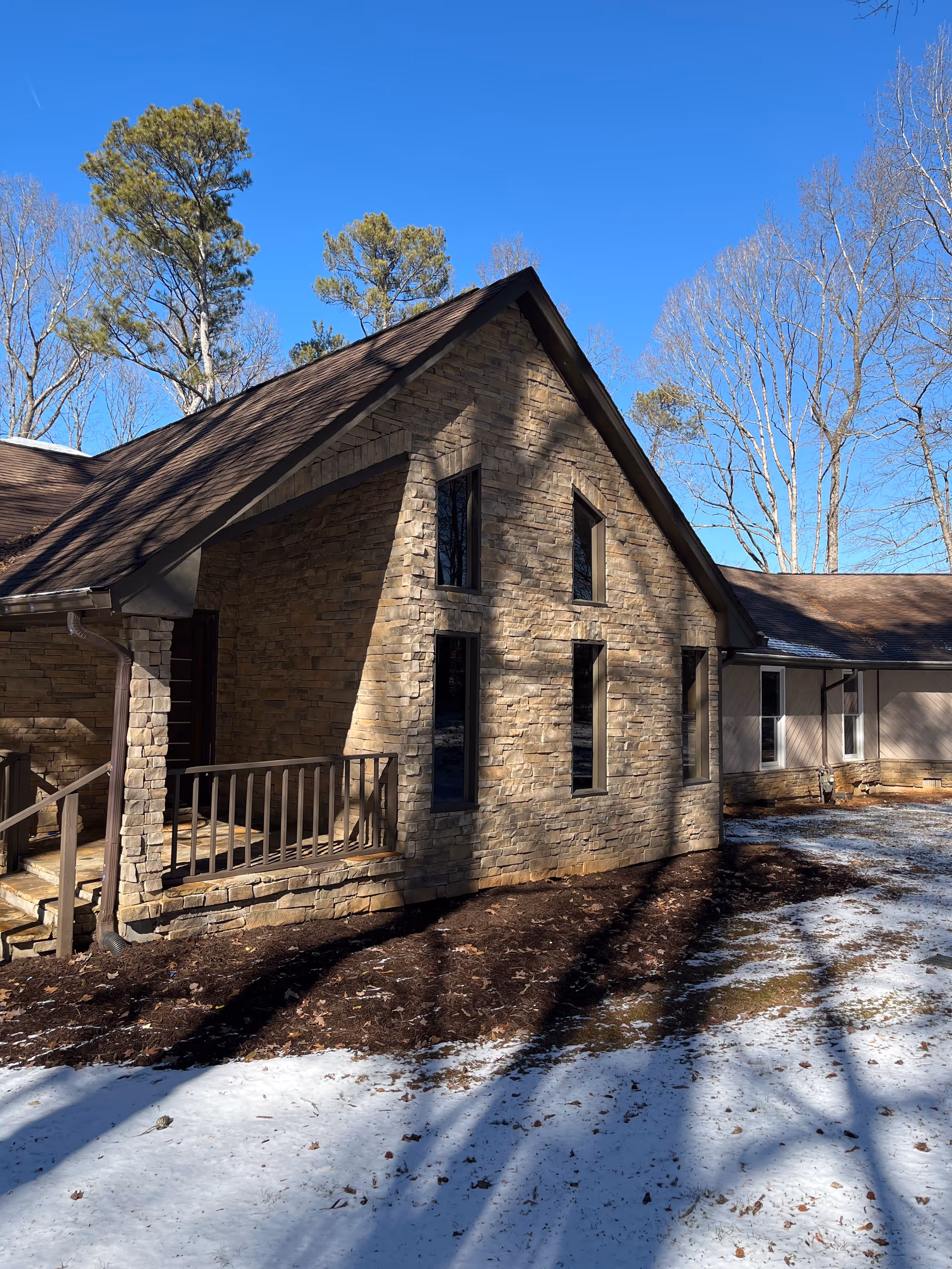 Exterior view of a senior living facility building with stone walls and a sloped roof. There is a small porch with wooden railings and steps leading up to the entrance. The ground is partially covered with snow and surrounded by leafless trees under a clear blue sky.