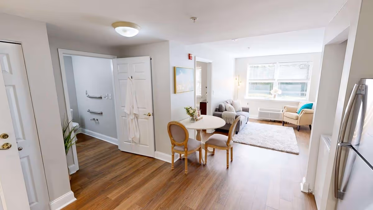 Bright and clean senior living apartment interior with wooden flooring, a small round dining table with two chairs, a gray sofa, an armchair near a large window, and a bathroom visible through an open door with grab bars and a white robe hanging on the door.