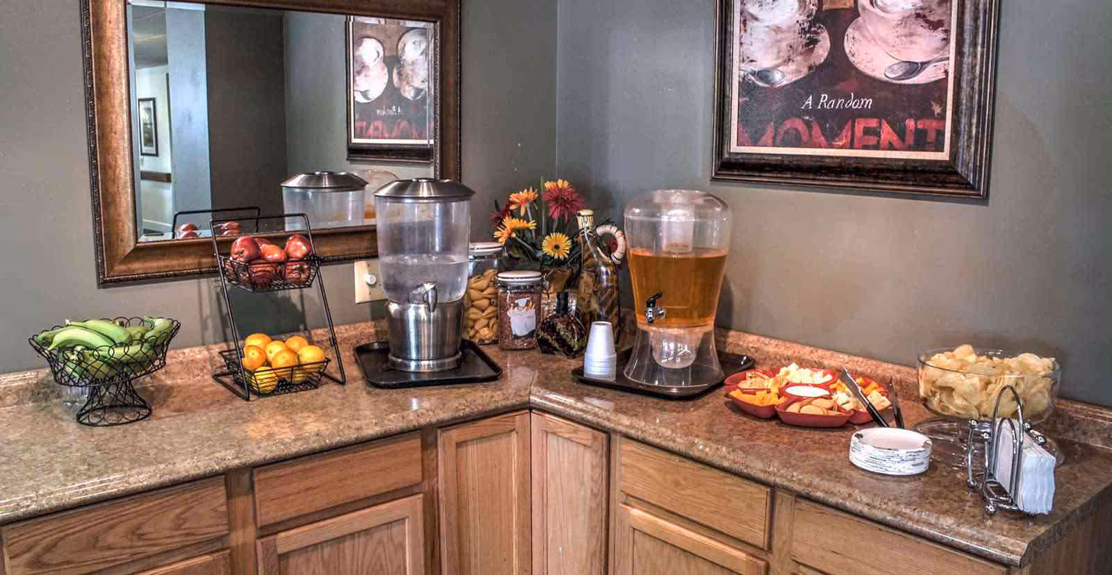 A countertop corner in a facility with a large mirror on the wall, featuring a water dispenser, a beverage dispenser with cups, baskets of bananas and apples, a tiered fruit basket with oranges, a bowl of chips, plates, napkins, and a framed artwork on the wall.