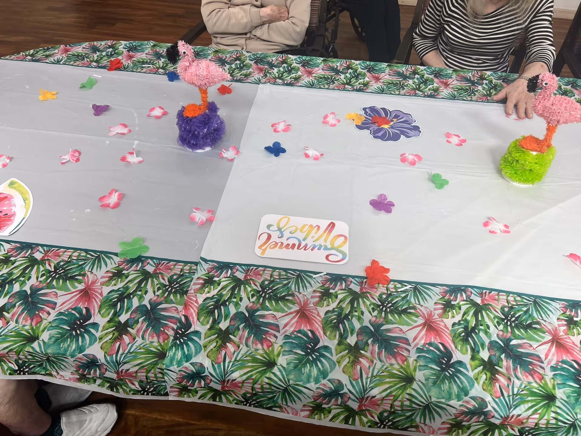 A table decorated with a tropical-themed tablecloth and a white plastic cover adorned with colorful flower and butterfly cutouts. Two small pink flamingo decorations are placed on the table. Three elderly individuals are partially visible sitting around the table.