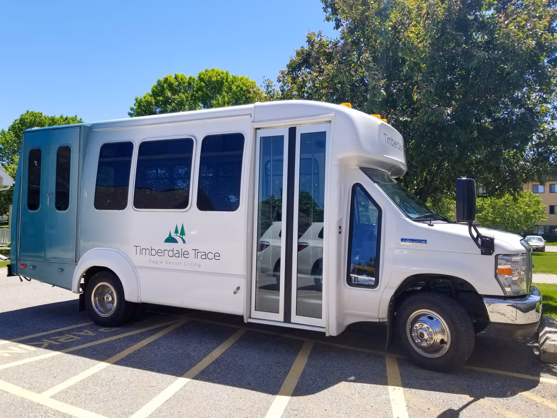 A white Timberdale Trace assisted living shuttle bus parked outdoors near trees and a building.