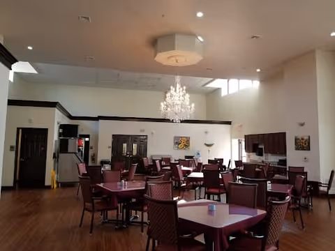A spacious dining room with multiple square tables and maroon cushioned chairs arranged neatly. The room features a large chandelier hanging from the ceiling, wood flooring, and high windows letting in natural light. There are paintings on the walls and a kitchenette area with cabinets and appliances in the background.