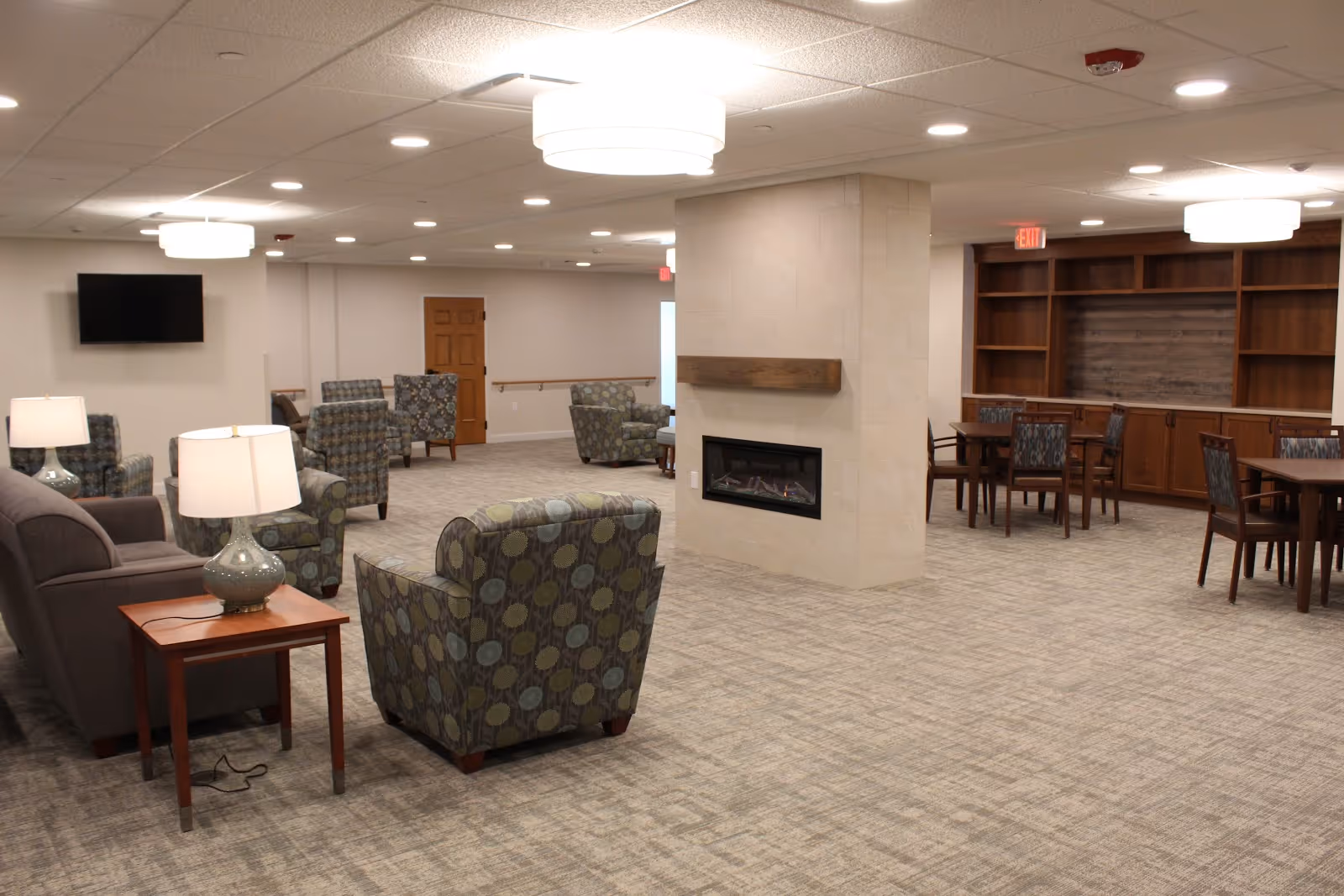 A spacious senior living common area with patterned armchairs, sofas, side tables with lamps, a wall-mounted TV, a central fireplace, and wooden shelving units with tables and chairs in the background. The room is well-lit with ceiling lights and has a neutral carpeted floor.