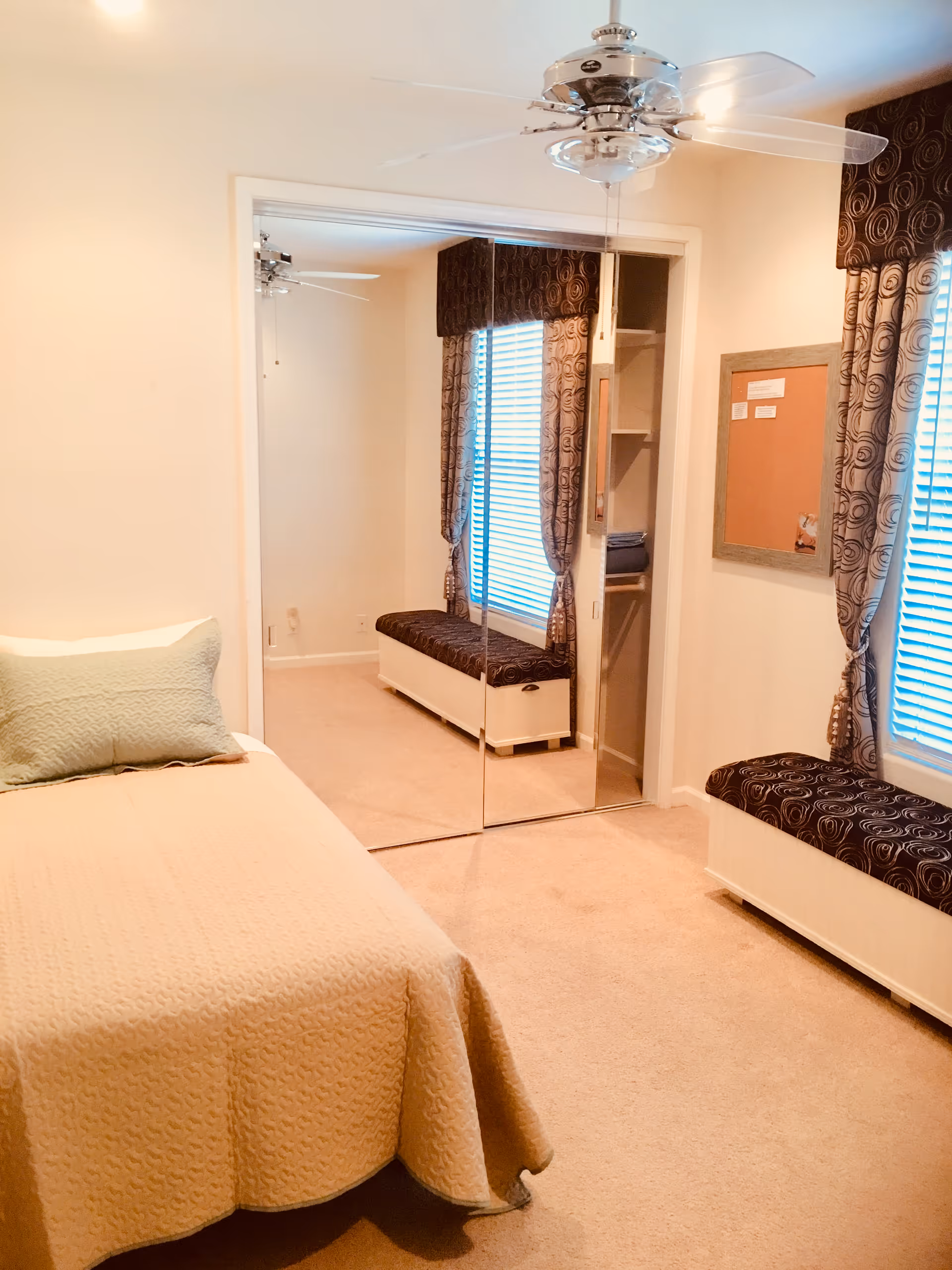 A cozy bedroom with a single bed covered in a beige quilt and a light green pillow. There is a window with patterned curtains and a cushioned bench underneath. A ceiling fan with lights is mounted on the ceiling. A mirrored closet door reflects the room, showing the bench and window from another angle. A bulletin board is mounted on the wall next to the window.