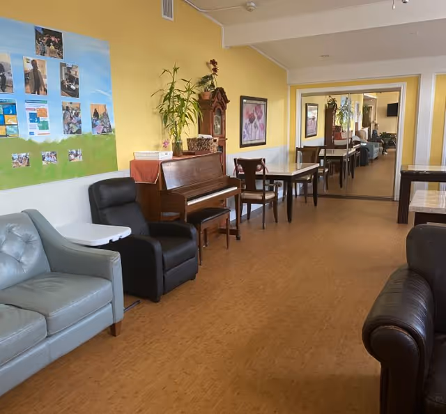 Communal lounge/dining area with sofas, a piano, tables and chairs along a yellow-painted wall and mirrored doorway.
