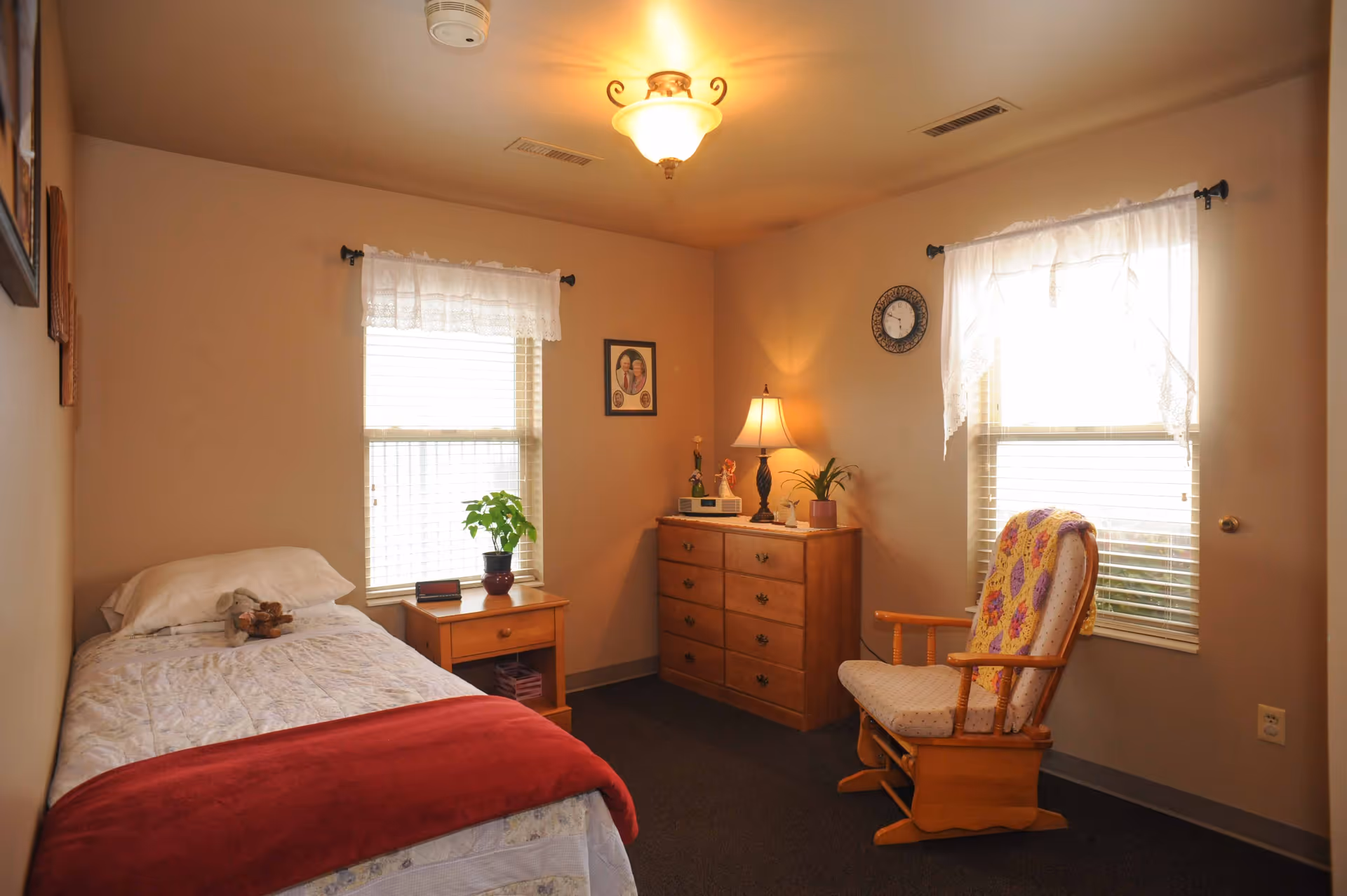 A cozy bedroom with a single bed covered in white bedding and a red blanket at the foot. A small wooden nightstand with a potted plant and a clock sits next to the bed. Across from the bed is a wooden dresser with a lamp, small plants, and decorative items. A cushioned wooden rocking chair with a colorful quilt is placed near a window with white lace curtains. The room has beige walls, carpeted floor, and a ceiling light fixture.