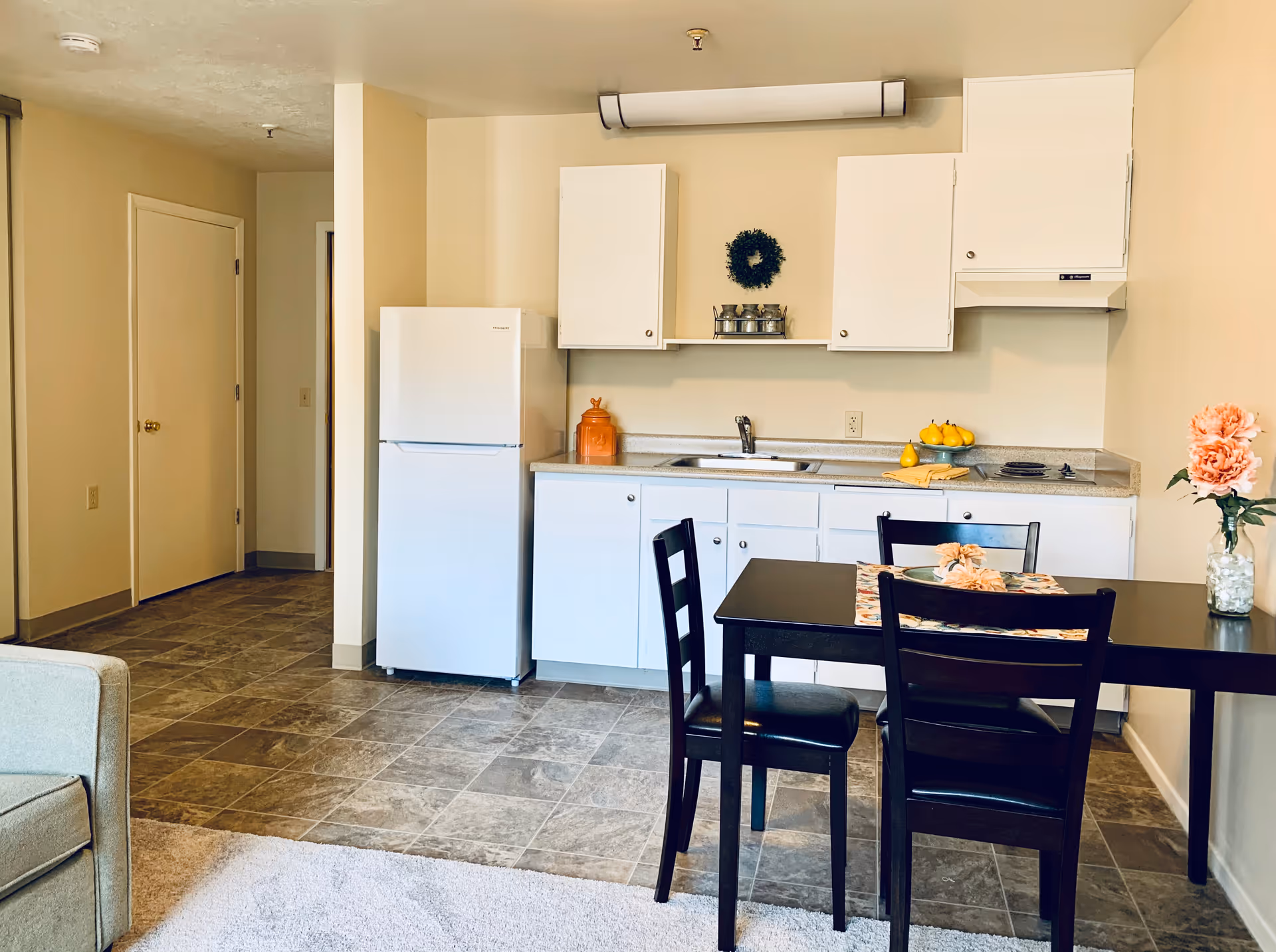 A small kitchen area with white cabinets, a white refrigerator, a sink, and a stove. There is a black dining table with three black chairs, decorated with a floral table runner and a flower centerpiece. The floor is tiled, and part of a beige carpet and a light-colored armchair are visible in the foreground. The walls are painted light beige, and there is a small green wreath and spice rack on the wall above the sink.