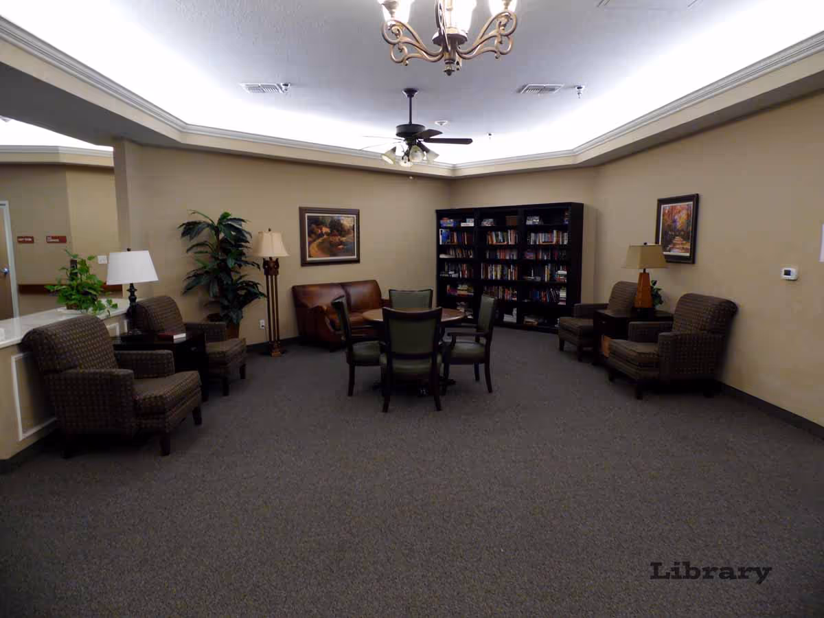 A cozy library room with a central round table surrounded by chairs, a brown leather couch, several armchairs, two floor lamps, a bookshelf filled with books, framed artwork on beige walls, a ceiling fan, and a chandelier.