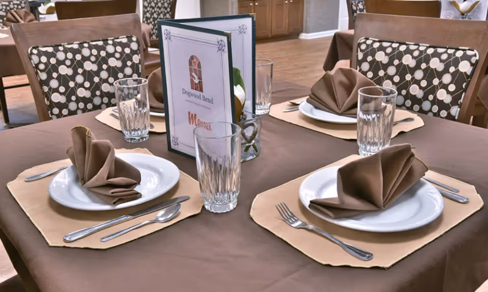 A dining table set for four with brown tablecloth and beige placemats, each place setting includes a white plate with a folded brown napkin, a fork, knife, spoon, and a clear drinking glass. In the center of the table is a menu card labeled 'Dogwood Bend' and a small glass vase with a white flower. The chairs have patterned cushions with a circular design.