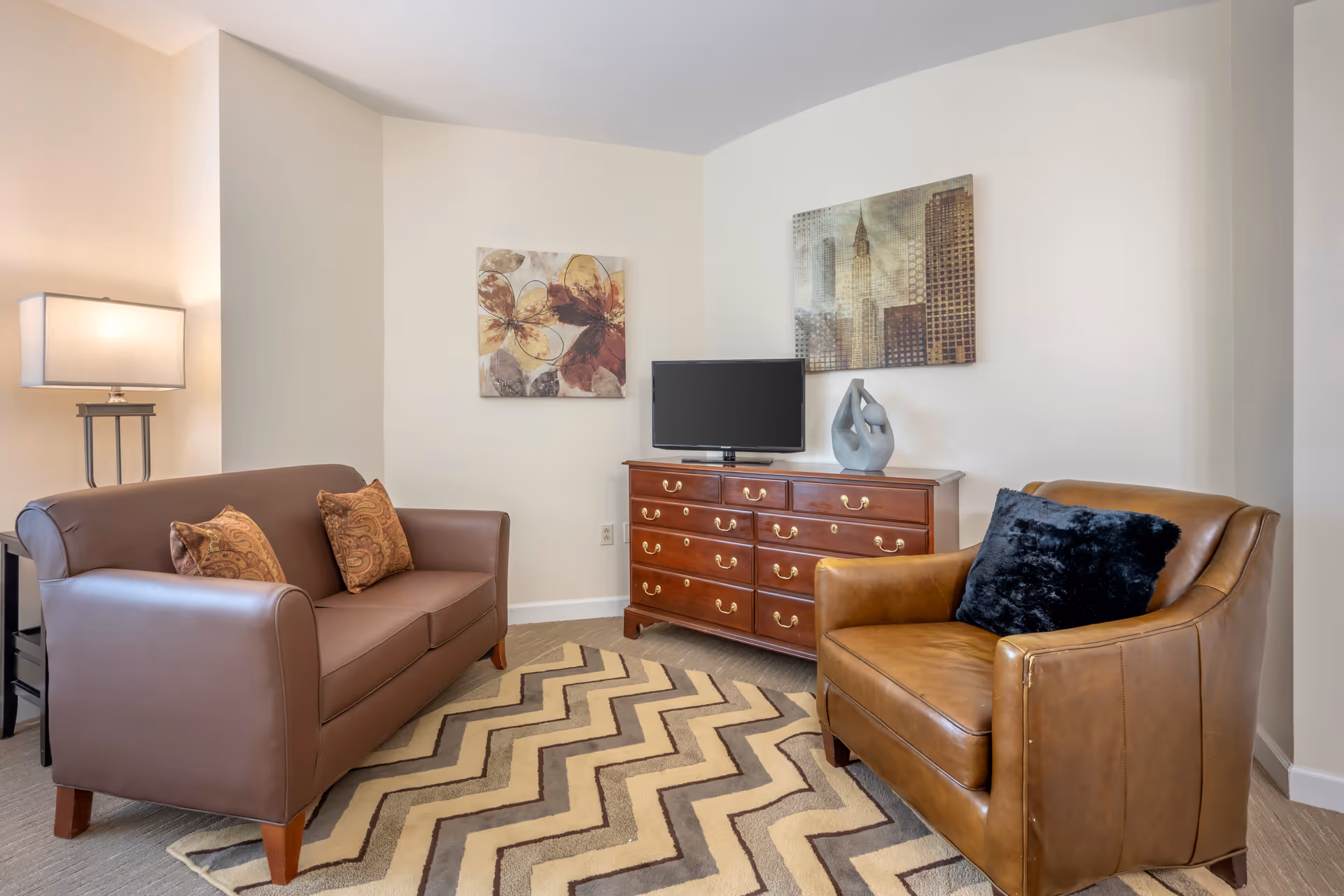 A cozy living room with a brown leather sofa with two patterned cushions, a tan leather armchair with a black cushion, a wooden dresser with a flat-screen TV on top, a modern sculpture, and two framed artworks on the wall. A floor lamp is positioned next to the sofa, and a beige and brown zigzag patterned rug covers the floor.