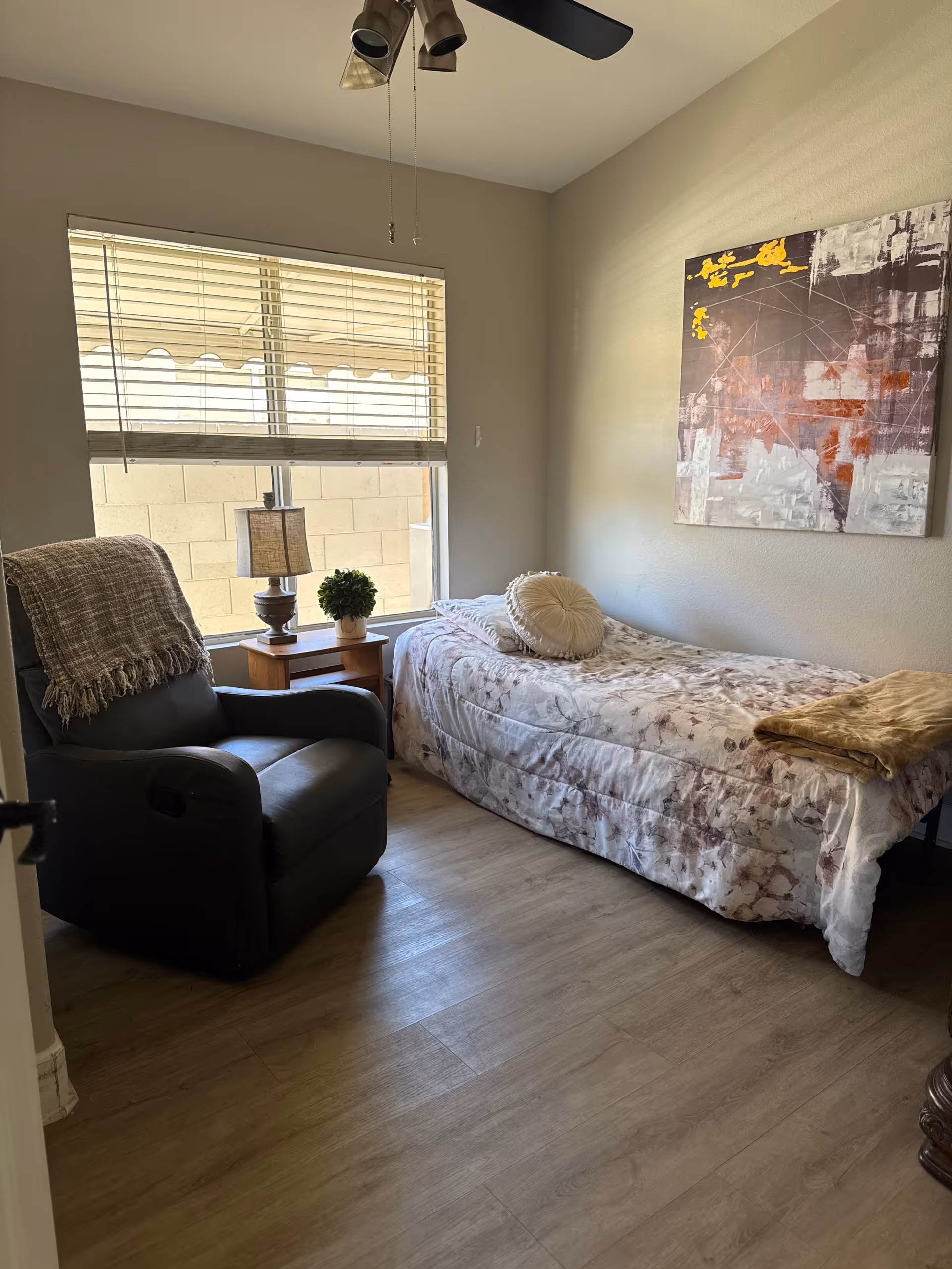 A small bedroom with a single bed covered in a patterned bedspread and a round decorative pillow. Next to the bed is a wooden side table with a lamp and a small potted plant. A dark recliner chair with a throw blanket draped over the back is positioned near a window with blinds. A ceiling fan with lights is visible above, and an abstract painting hangs on the wall above the bed.