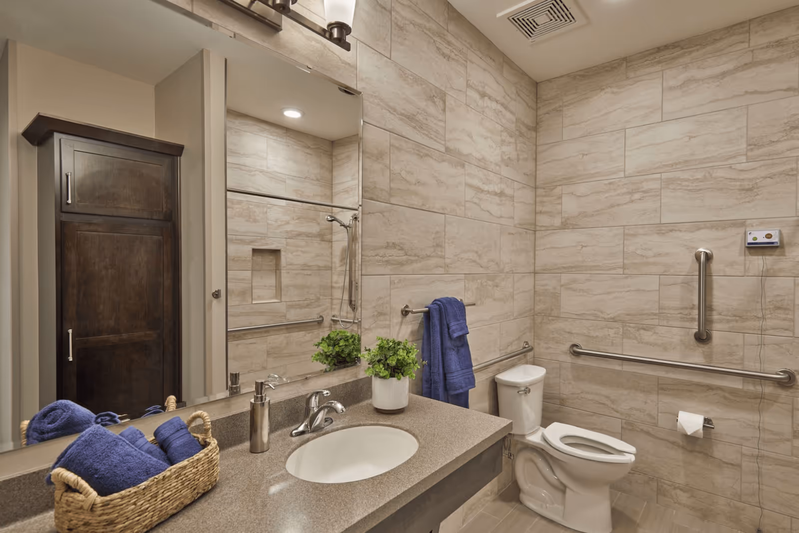 A clean and modern bathroom with beige tiled walls and floor. The bathroom features a white toilet with grab bars on the walls for accessibility, a countertop with an oval sink, a soap dispenser, a small green potted plant, and a basket with neatly folded blue towels. A large mirror is mounted above the sink, reflecting part of the shower area with a handheld showerhead and a built-in niche. A dark wooden cabinet is visible in the reflection.