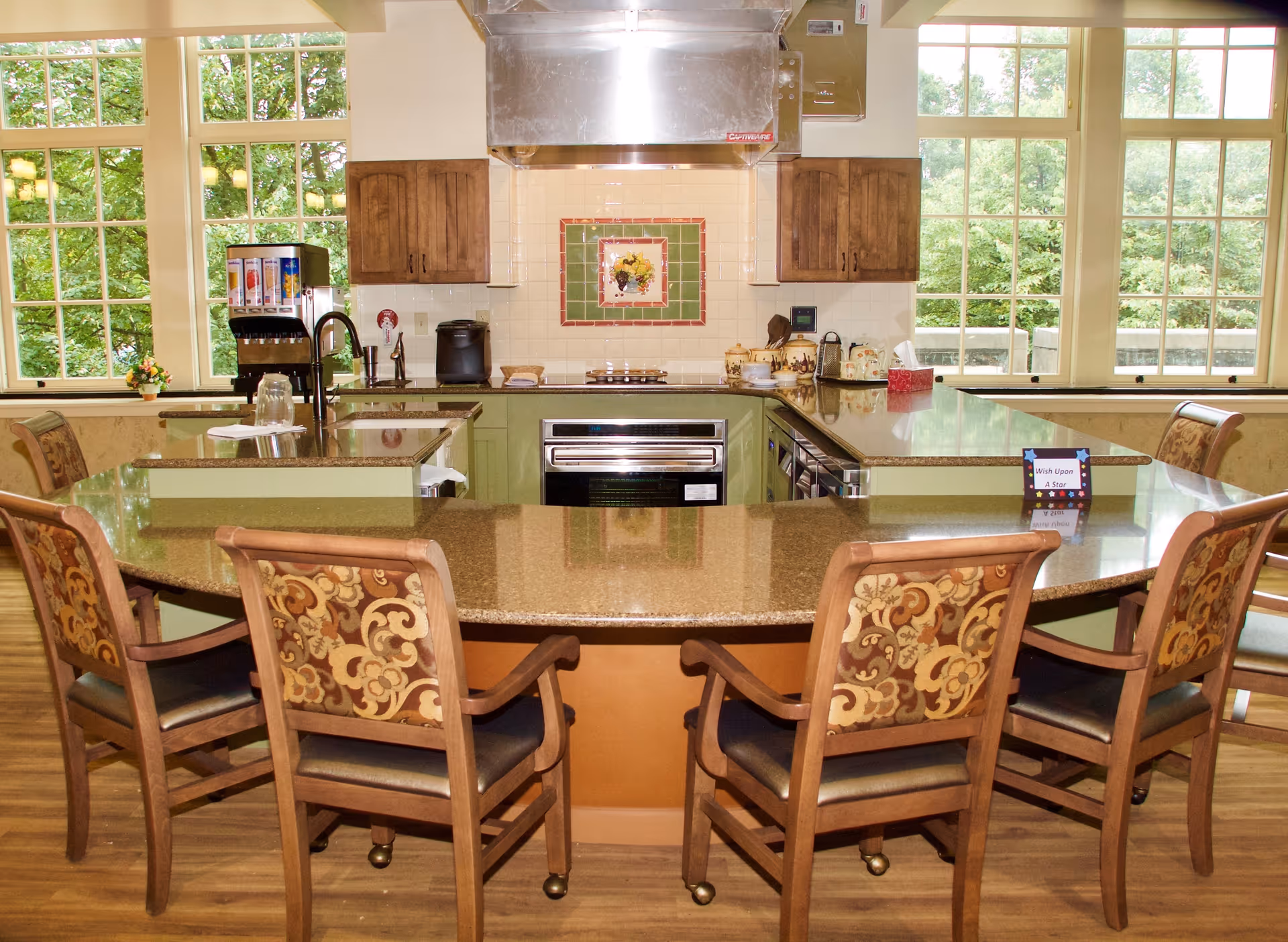A bright kitchen area with a large U-shaped granite countertop surrounded by six wooden chairs with patterned upholstery. The kitchen features green lower cabinets, a stainless steel oven, a stove with a stainless steel hood, and large windows showing green trees outside. Various kitchen appliances and decorative items are placed on the countertops.