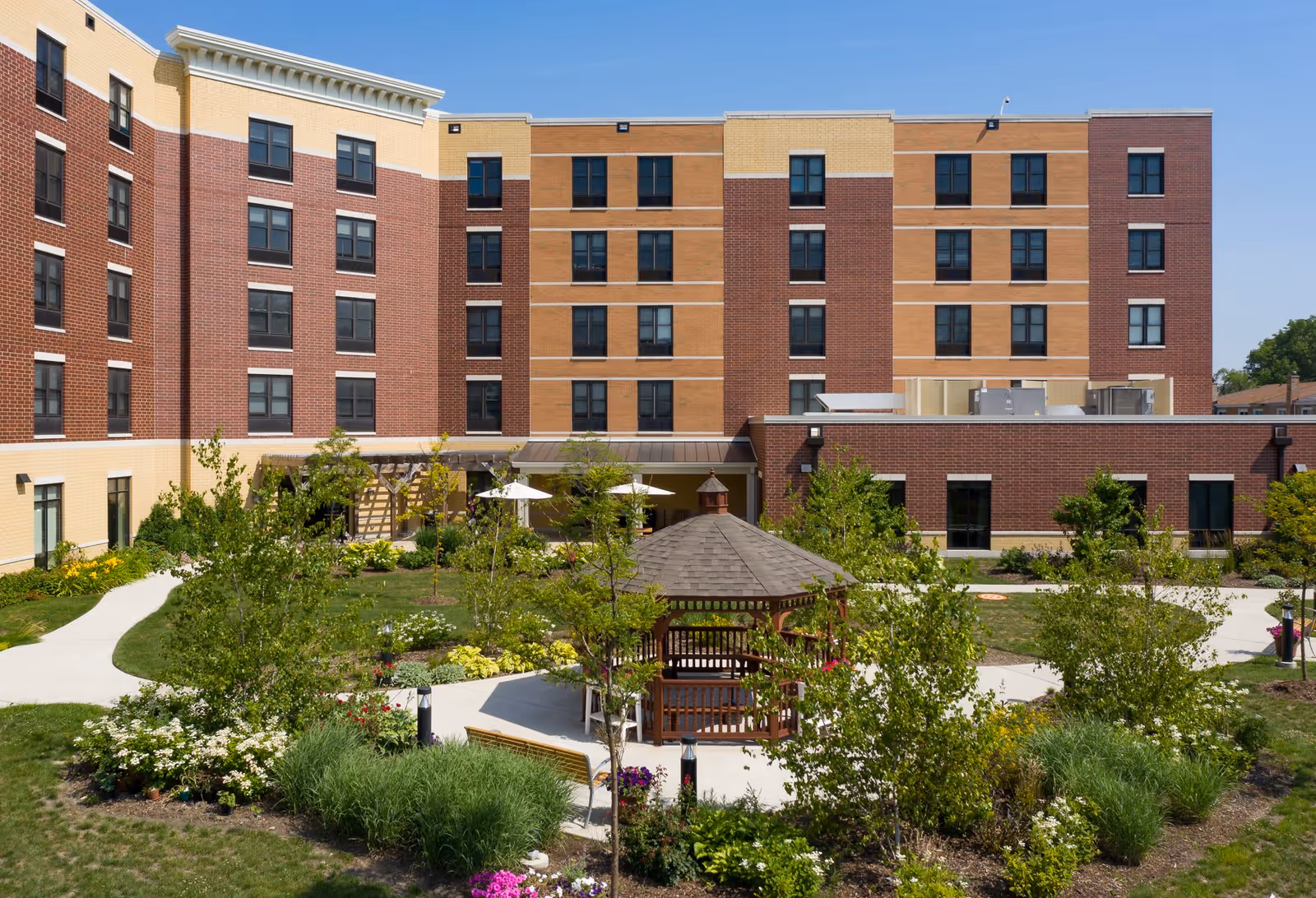Outdoor garden area at Montclare Supportive Living Community featuring a wooden gazebo surrounded by greenery, flowers, and walking paths, with a multi-story brick and tan building in the background under a clear blue sky.