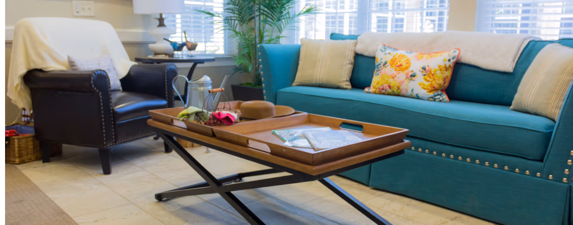 A cozy living room with a teal sofa adorned with floral and beige pillows, a dark leather armchair with a white throw draped over it, and a wooden coffee table with a tray holding a hat, flowers, and some papers. The room has large windows with white blinds and a potted plant in the corner.
