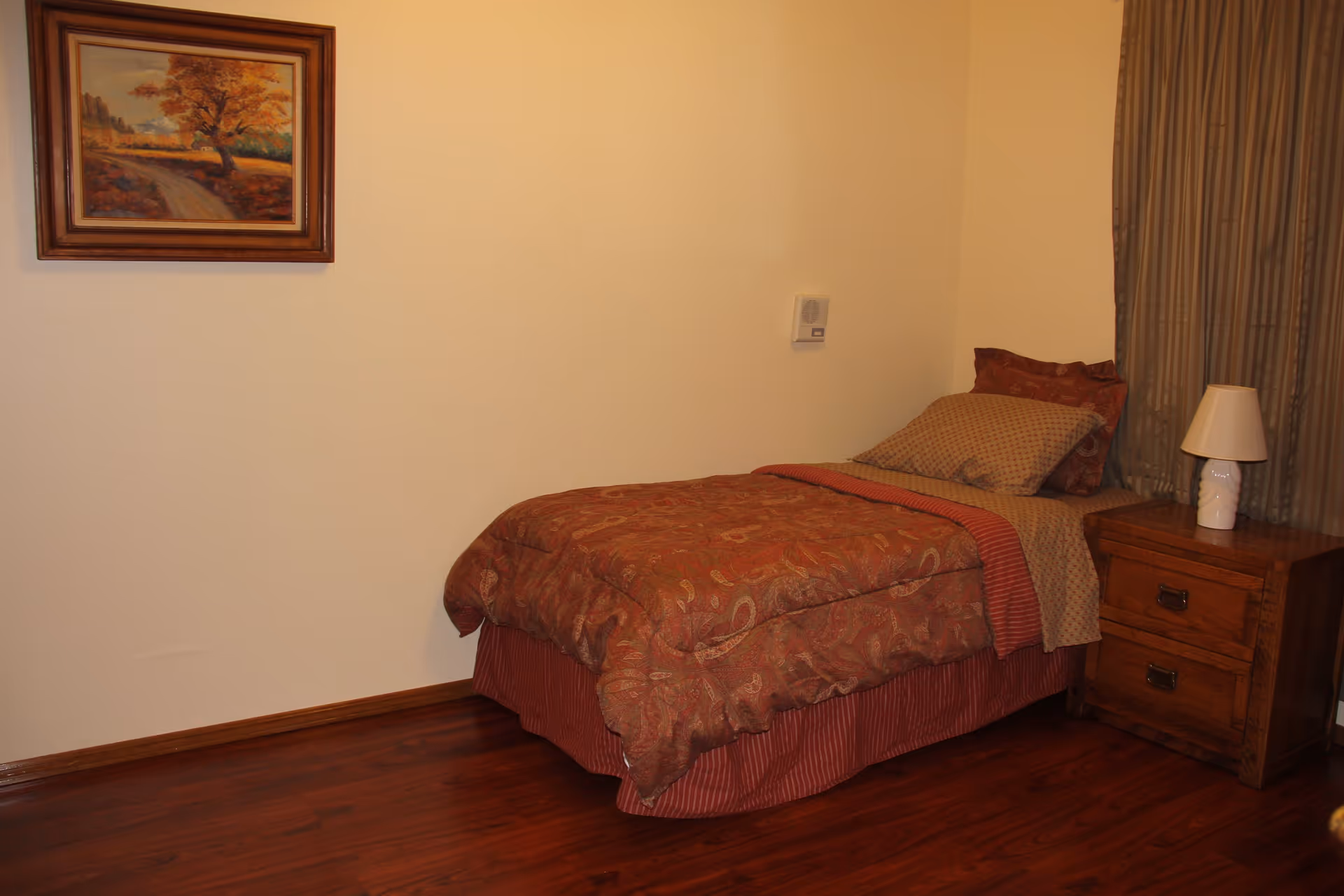 A small bedroom with a single bed covered in a patterned red and brown comforter and pillows. Next to the bed is a wooden nightstand with two drawers and a white lamp on top. The room has wooden flooring, beige walls, a framed painting of a tree on the wall, and brown curtains covering a window.