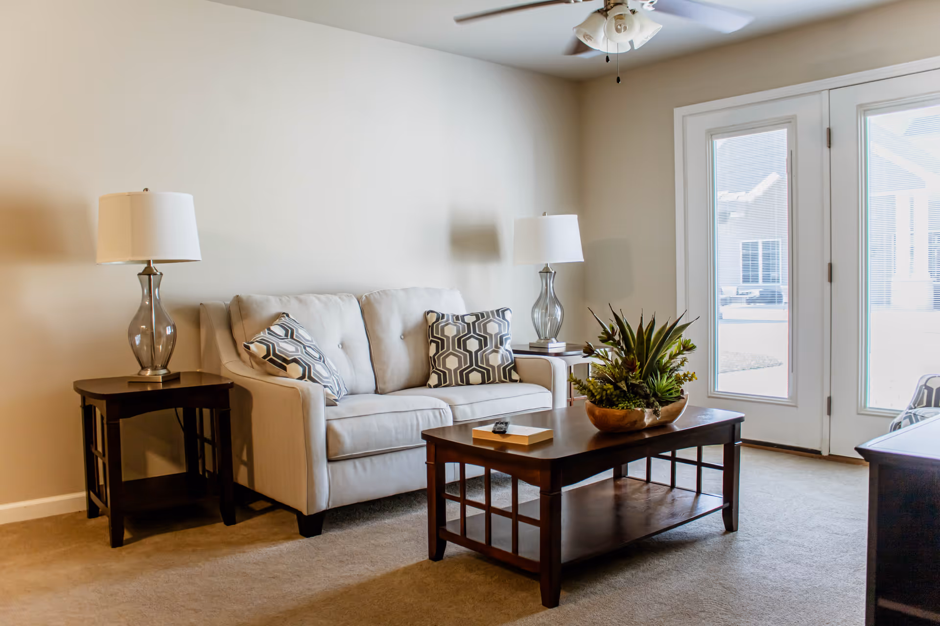 Bright living room with a beige sofa, patterned pillows, a wooden coffee table with a plant, side tables with lamps, and glass doors to the outside.