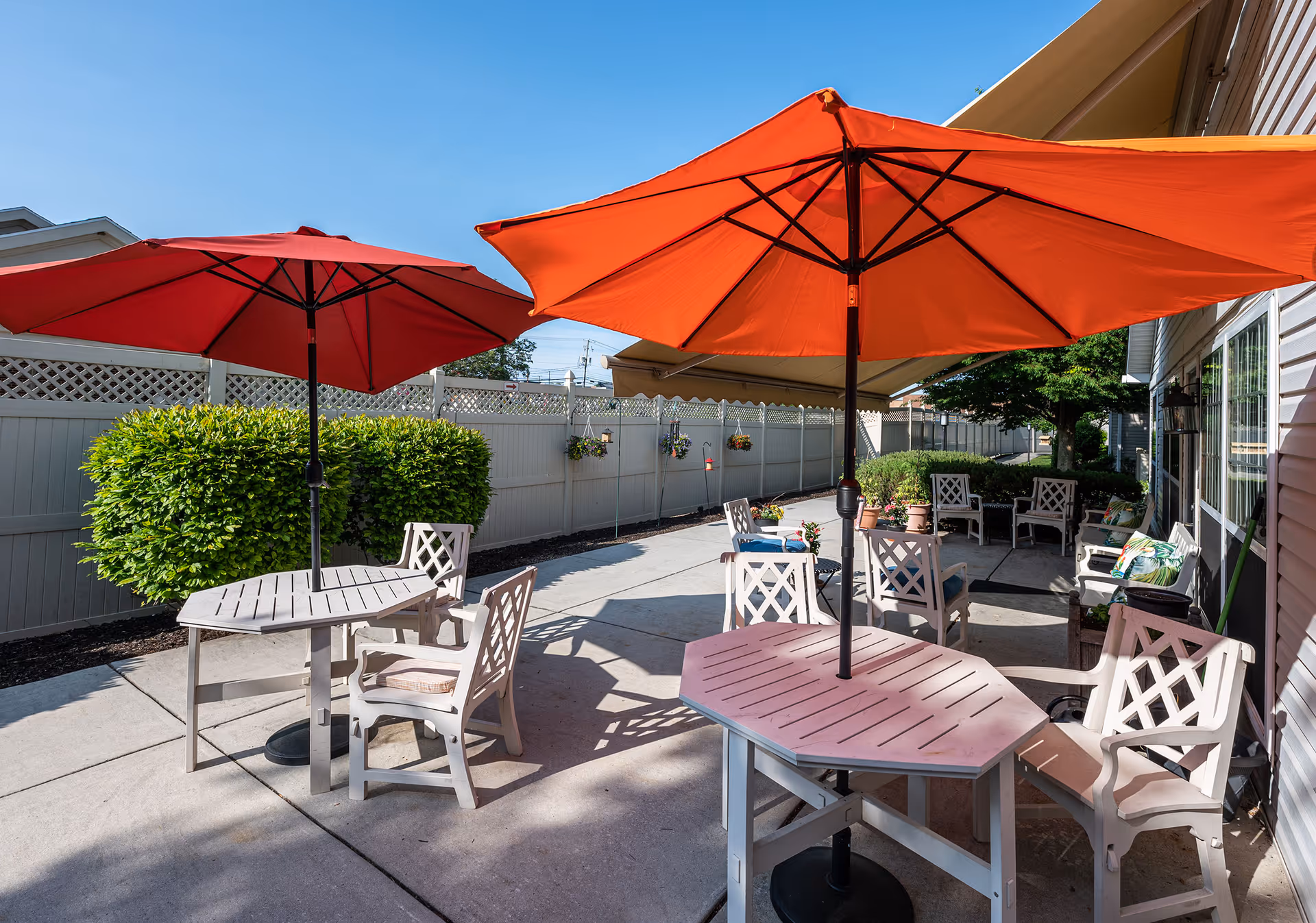 Outdoor patio area with several white tables and chairs, each table shaded by large red umbrellas. The patio is surrounded by a white fence and green bushes, with a clear blue sky overhead.