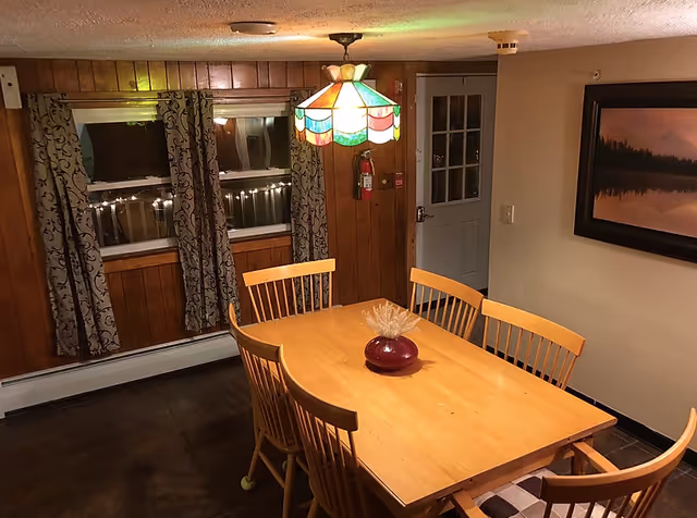 A dining room with a wooden table surrounded by six wooden chairs. A colorful stained glass pendant light hangs above the table. The room has wood-paneled walls with two windows covered by patterned curtains. A fire extinguisher is mounted on the wall near a door with glass panes. A framed landscape picture hangs on the adjacent wall.
