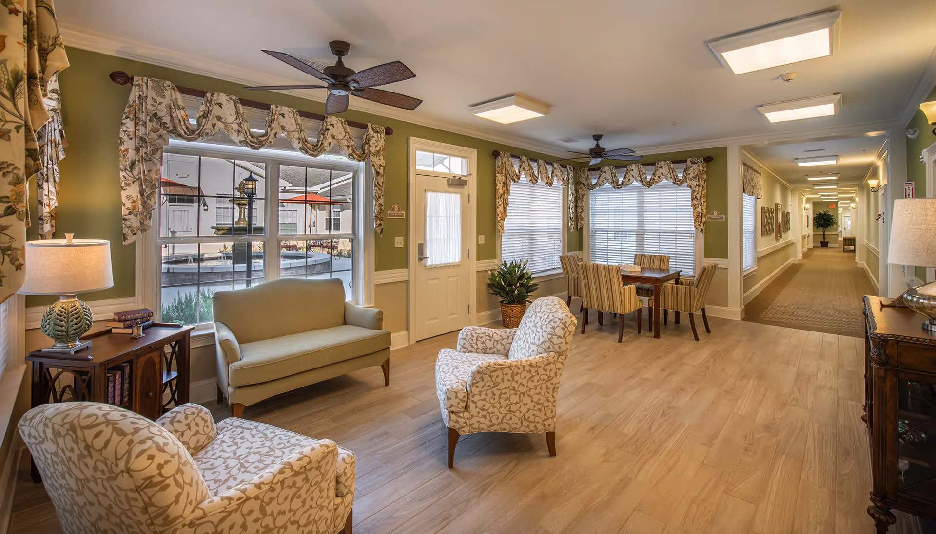 Sunlit common seating area with armchairs, a loveseat, dining table, and large windows in a senior living facility.