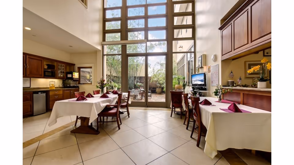 Bright double-height dining area with tables covered in white linens and purple napkins, wooden chairs, a kitchenette, and floor-to-ceiling windows opening to a courtyard.