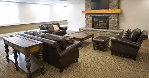 A cozy living room area with brown leather sofas, a matching ottoman, a wooden coffee table, and a side table. The room features a stone fireplace with a wooden mantel and a mounted flat-screen TV above it. The floor is carpeted with a patterned design, and there are large windows with white blinds letting in natural light.