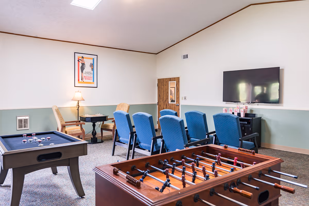 A recreational room with a foosball table in the foreground, a shuffleboard table to the left, and a row of blue chairs facing a wall-mounted flat screen TV. There are two armchairs with a small table and lamp between them, and a framed poster on the wall. The room has light green and white walls and a carpeted floor.