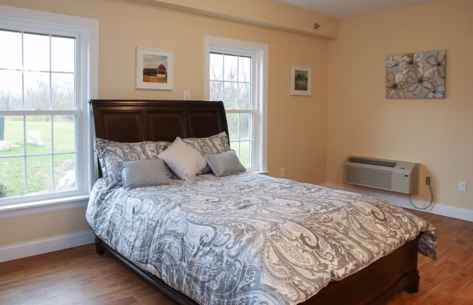 A bedroom with a large wooden bed frame and a paisley patterned bedspread in shades of gray and white. There are three pillows on the bed, two gray and one beige. The room has light beige walls, two large windows showing a green outdoor area, a wall-mounted air conditioning unit, and three pieces of wall art including a floral painting and two smaller framed pictures.